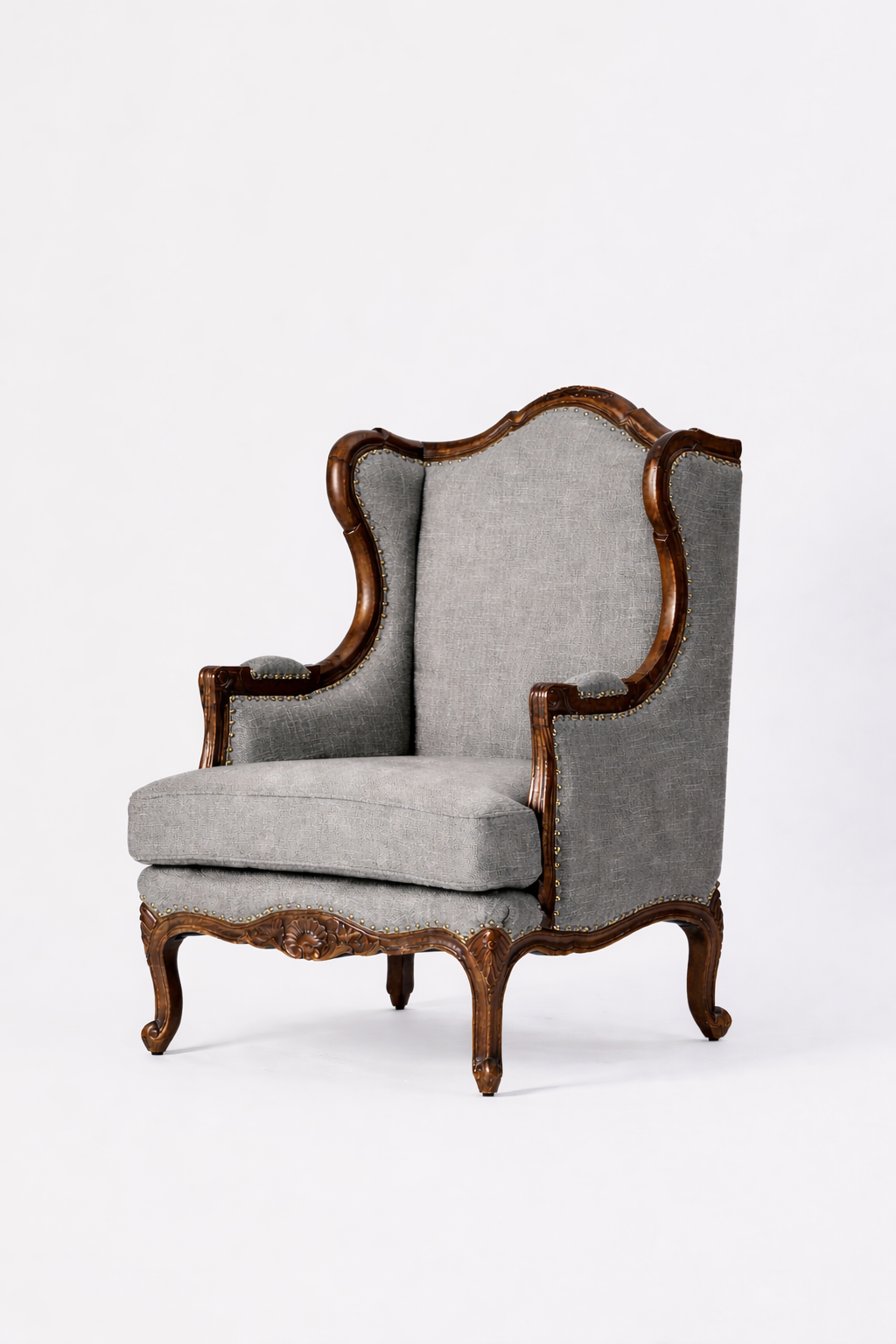 The Montclair Heritage Wingback Armchair features gray upholstered cushions, a high back, handcrafted solid wood frame with ornate carvings, and curved legs, displayed against a plain white background.