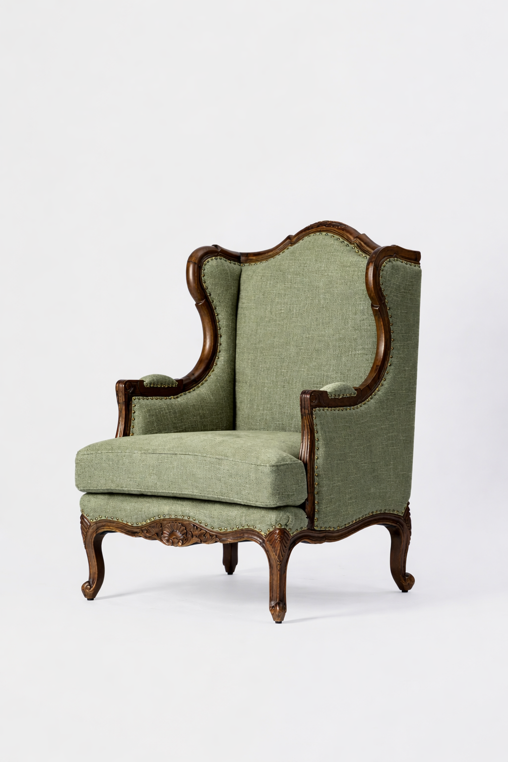 The Montclair Heritage Wingback Armchair features a green upholstered seat and backrest with ornate, handcrafted solid wood carvings on its arms, legs, and frame, showcased against a plain white background.