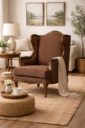 A cozy living room featuring the Montclair Heritage Wingback Armchair in brown, draped with a beige throw, a round woven ottoman with a teacup set, a solid wood side table with lamp and flowers, and two landscape paintings on the wall.
